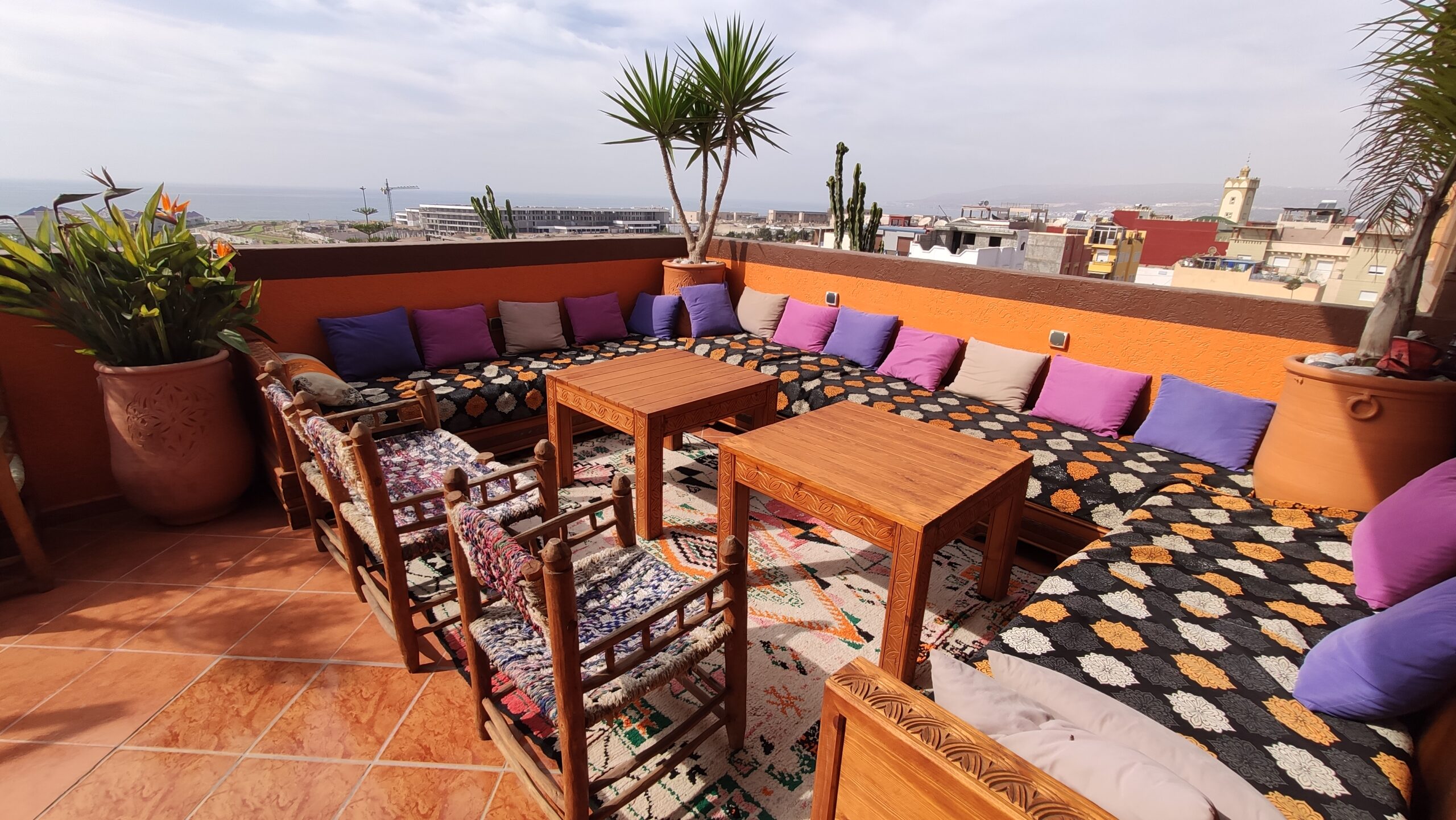 Terrasse im Azul Guesthouse in Tamraght, Marokko, mit bequemen Sitzgelegenheiten, üppigem Grün und einem atemberaubenden Blick auf den Ozean und die umliegenden Berge, ideal für Surf- und Yoga-Aufenthalte.