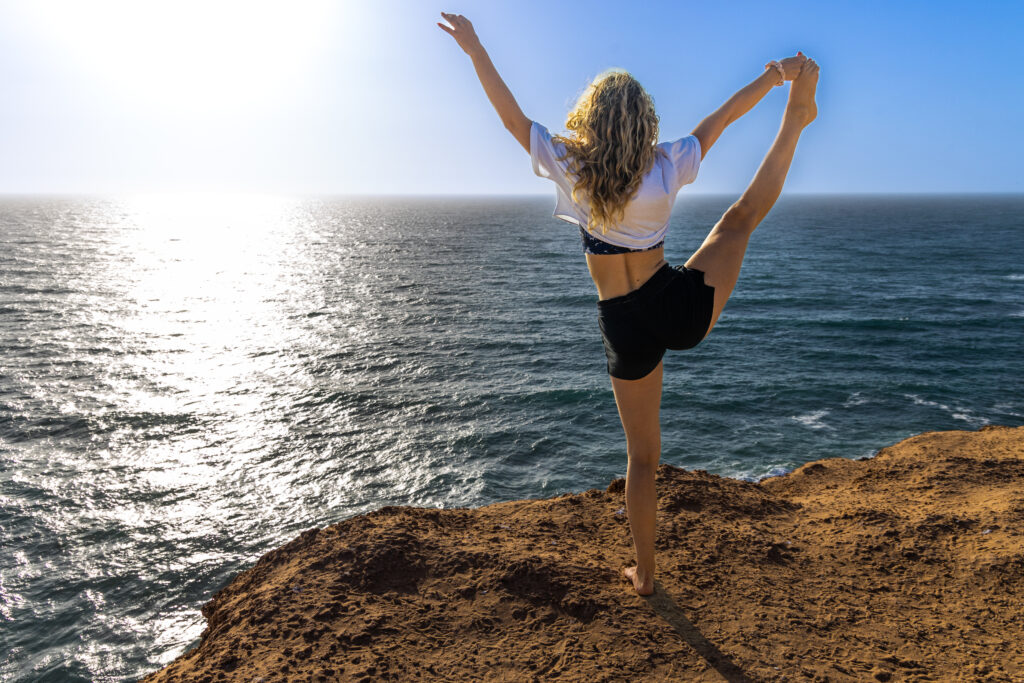 Eine Frau führt eine Yogapose an einem goldenen Sandstrand in Timlalin aus, mit dem Ozean im Hintergrund und der untergehenden Sonne.